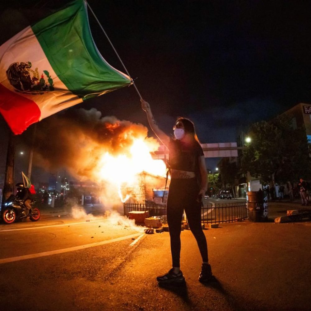 A woman waves the Mexican flag as flames erupt from a burning dumpster during a protest in downtown Los Angeles, Sunday, June 8, 2025. (AP Photo/Ethan Swope) Alabama News