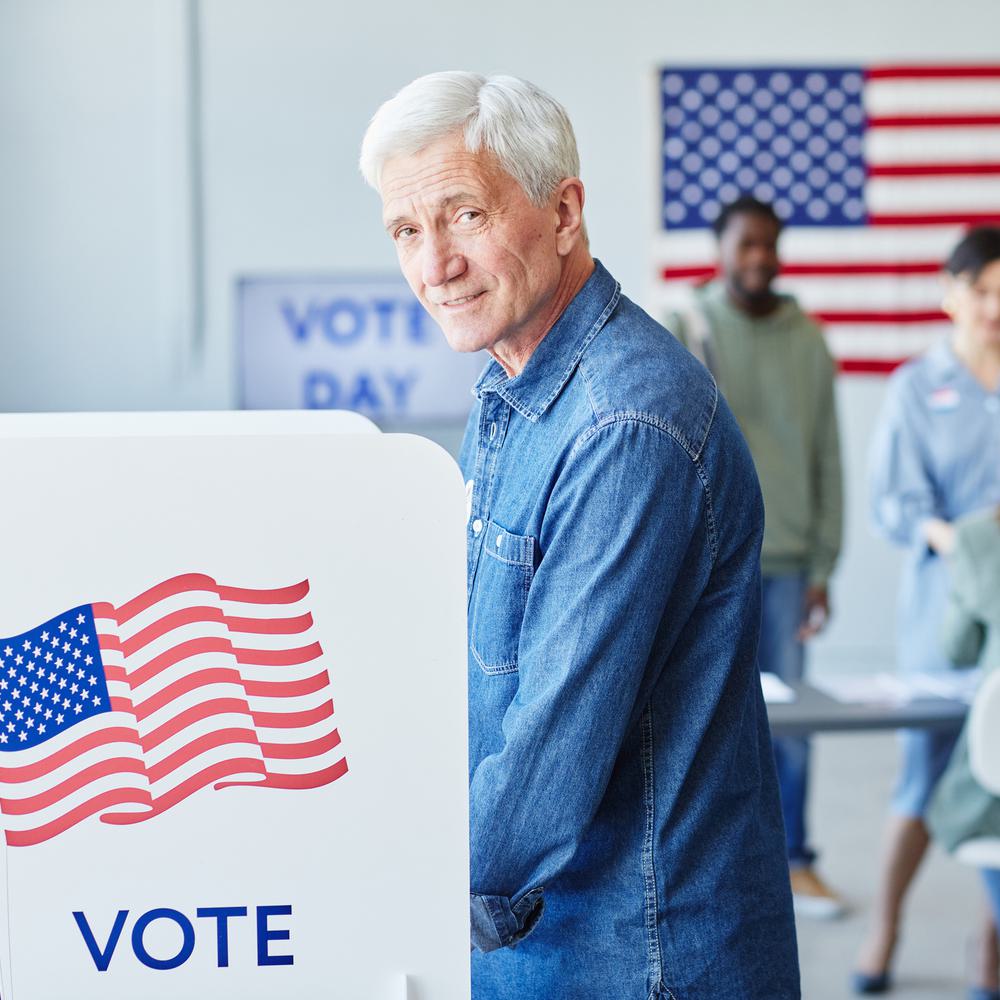 Side view portrait of smiling senior man voting in booth on election day and looking at camera, copy space Alabama News